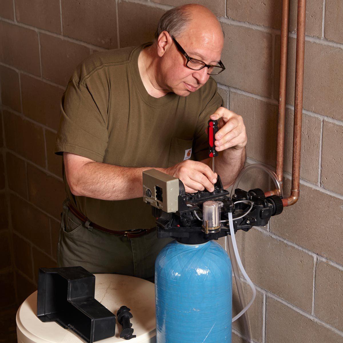 A man repairs a device, using a screwdriver, on a blue tank while positioned at a table in a gray, concrete-walled environment.