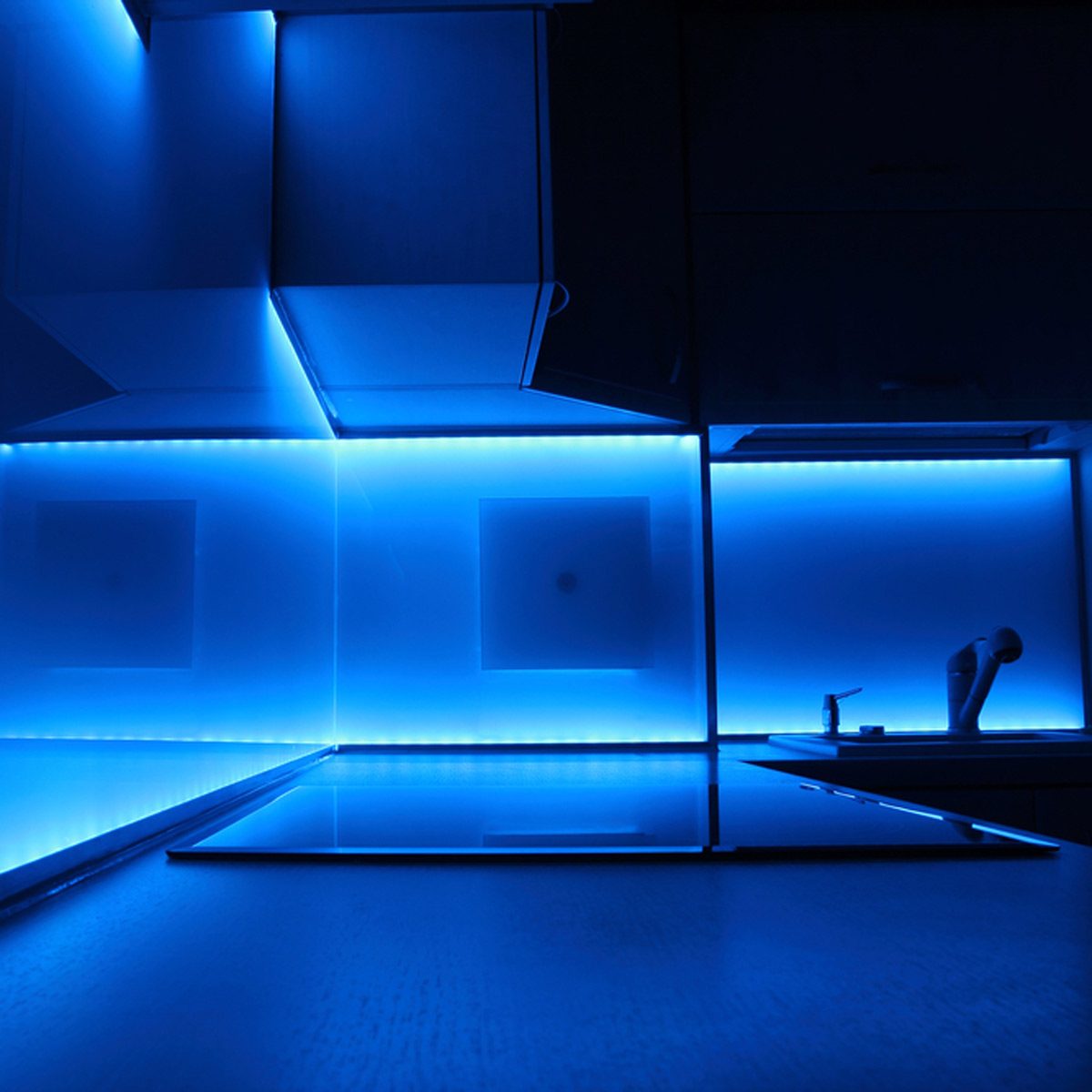 A modern kitchen features sleek surfaces illuminated by vibrant blue LED lights, creating a serene and stylish atmosphere. Countertops reflect the colorful glow.