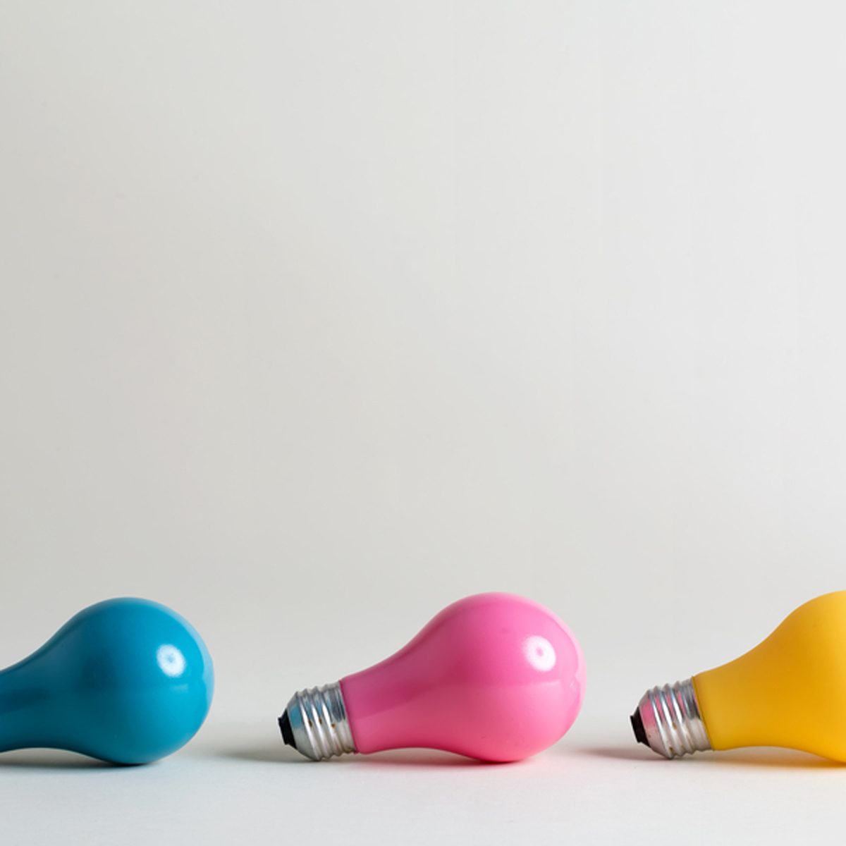 Three colorful light bulbs—blue, pink, and yellow—are positioned on a light gray surface against a plain, soft background.