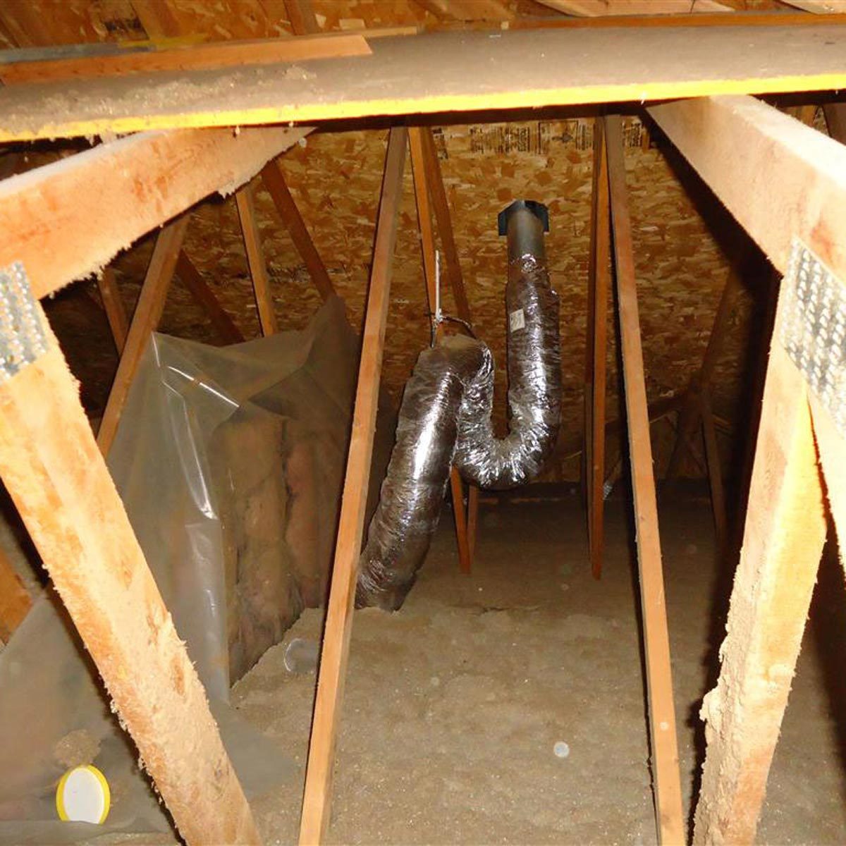 An insulated duct runs horizontally through a dimly lit attic, surrounded by wooden beams and insulation material, with some debris on the ground.