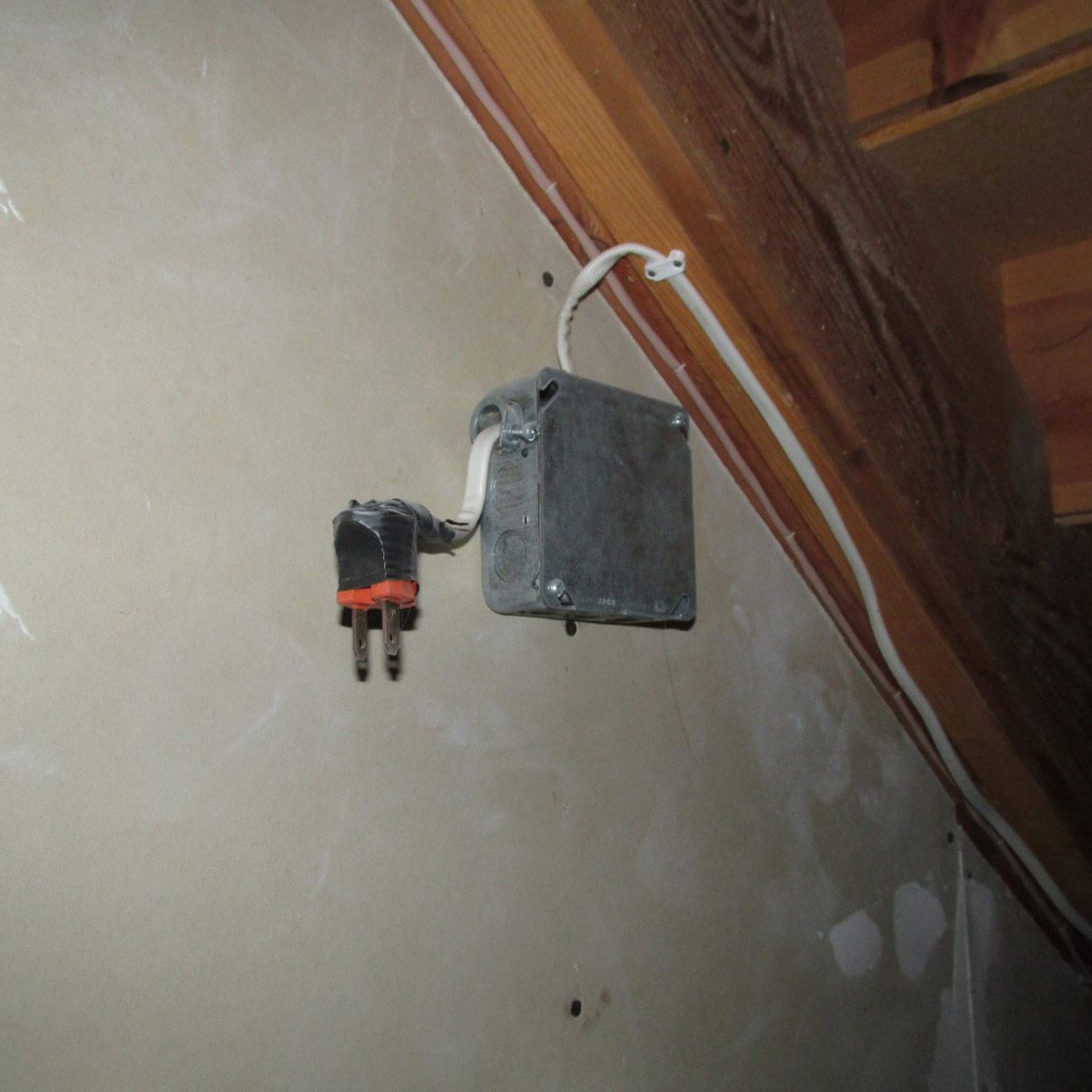 A metal electrical junction box is mounted on a wall, connected to a black and orange plug, with a white cable extending to the ceiling rafters.