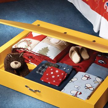 A yellow storage drawer is open, revealing neatly folded blankets and clothing, alongside a stuffed bear, in a bedroom with a colorful quilted bedspread above.