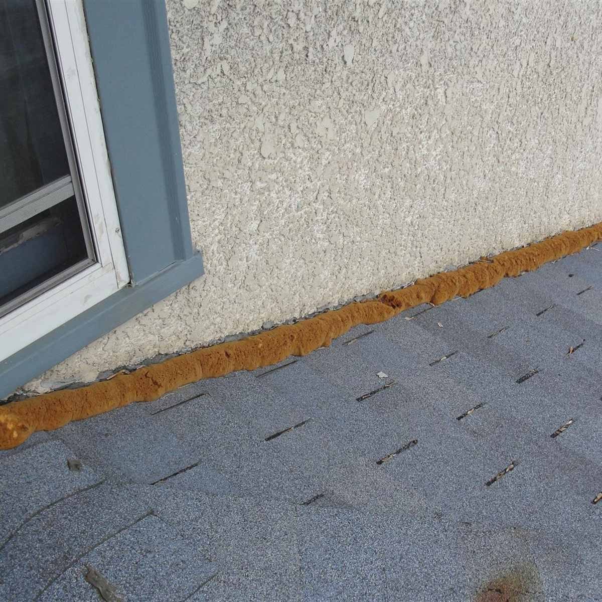 A brown, fuzzy material lines the edge of a gray roof, adjacent to a textured beige wall and a nearby window.