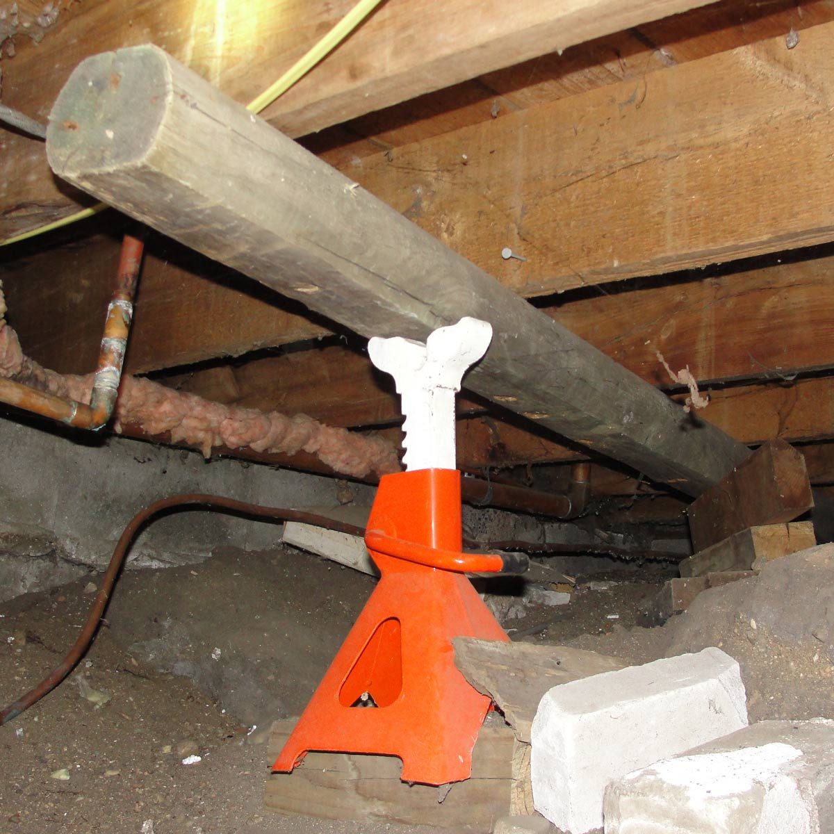 A wooden beam rests on a bright orange jack, stabilizing it in a dimly lit basement with exposed pipes and dirt flooring.
