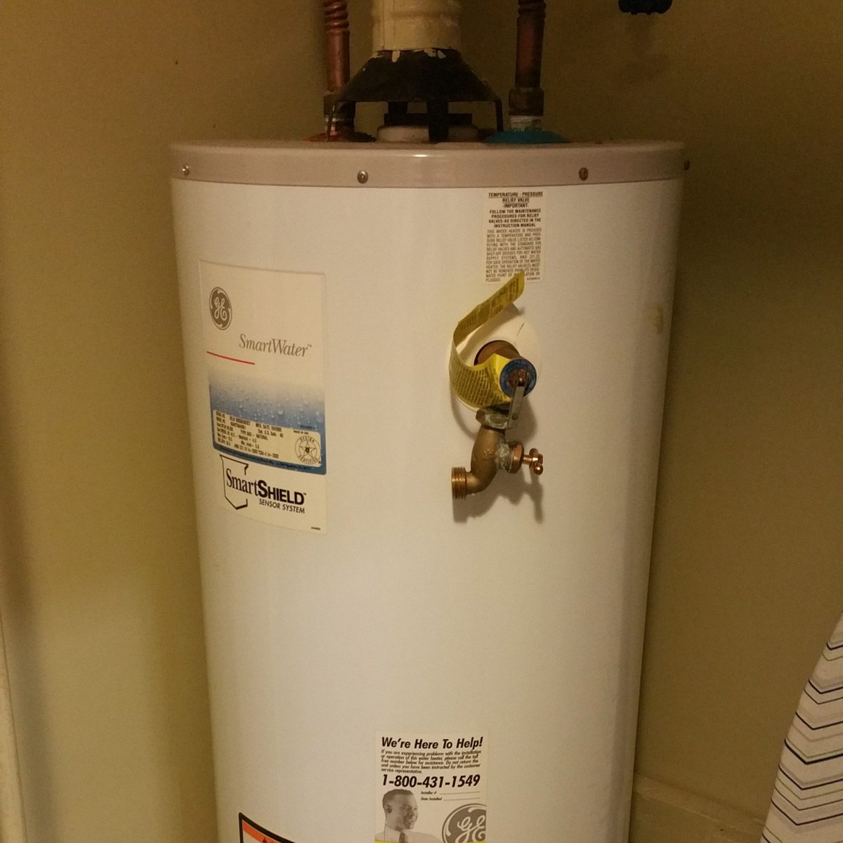 A cylindrical water heater stands against a wall, featuring a valve and control system, with instructional labels and contact information attached.