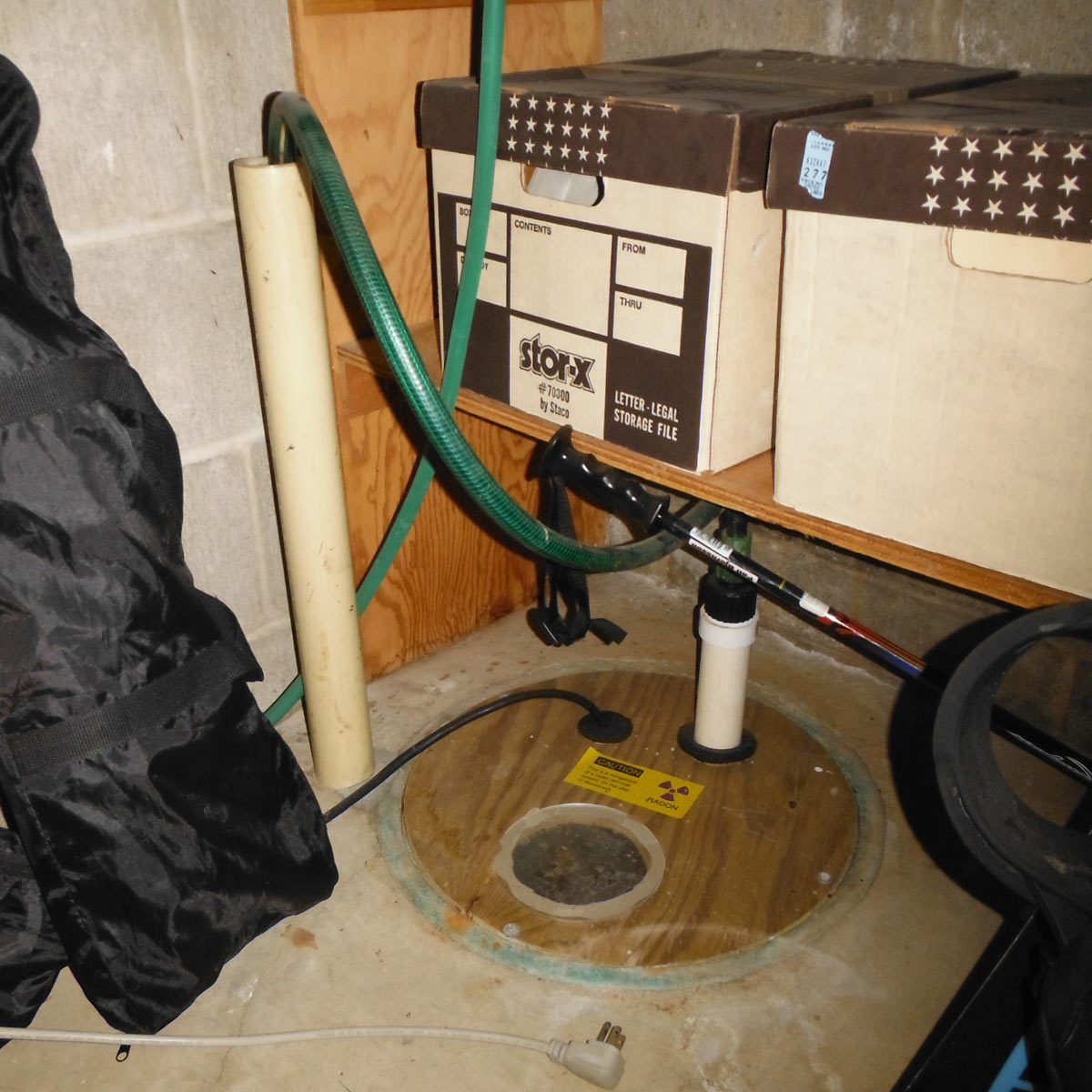 A sump pump is positioned on a wooden platform, connected to a hose, beside storage boxes in a dimly lit basement.