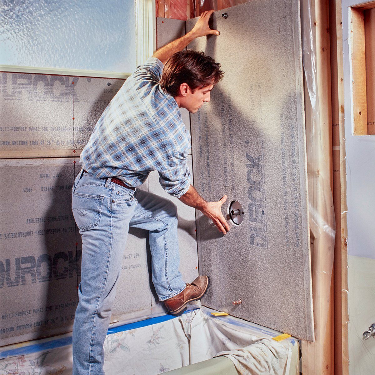 A man installs a wall panel in a partially finished room, using his foot for support and aligning it around a pipe fitting.