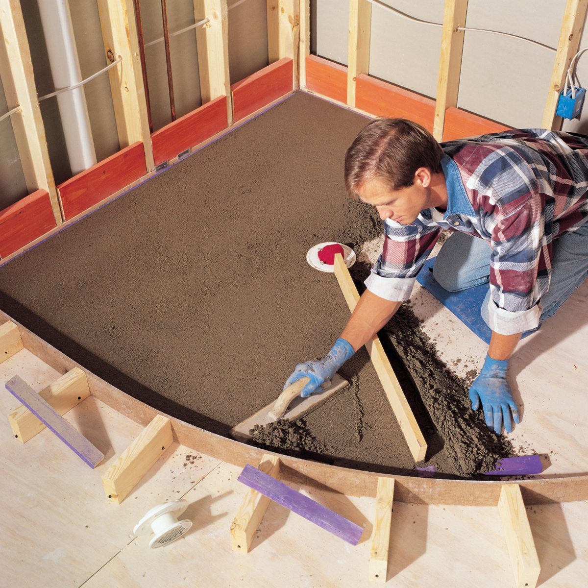 A man is leveling wet concrete using a trowel inside a partially constructed room with wooden framing and a curved edge.