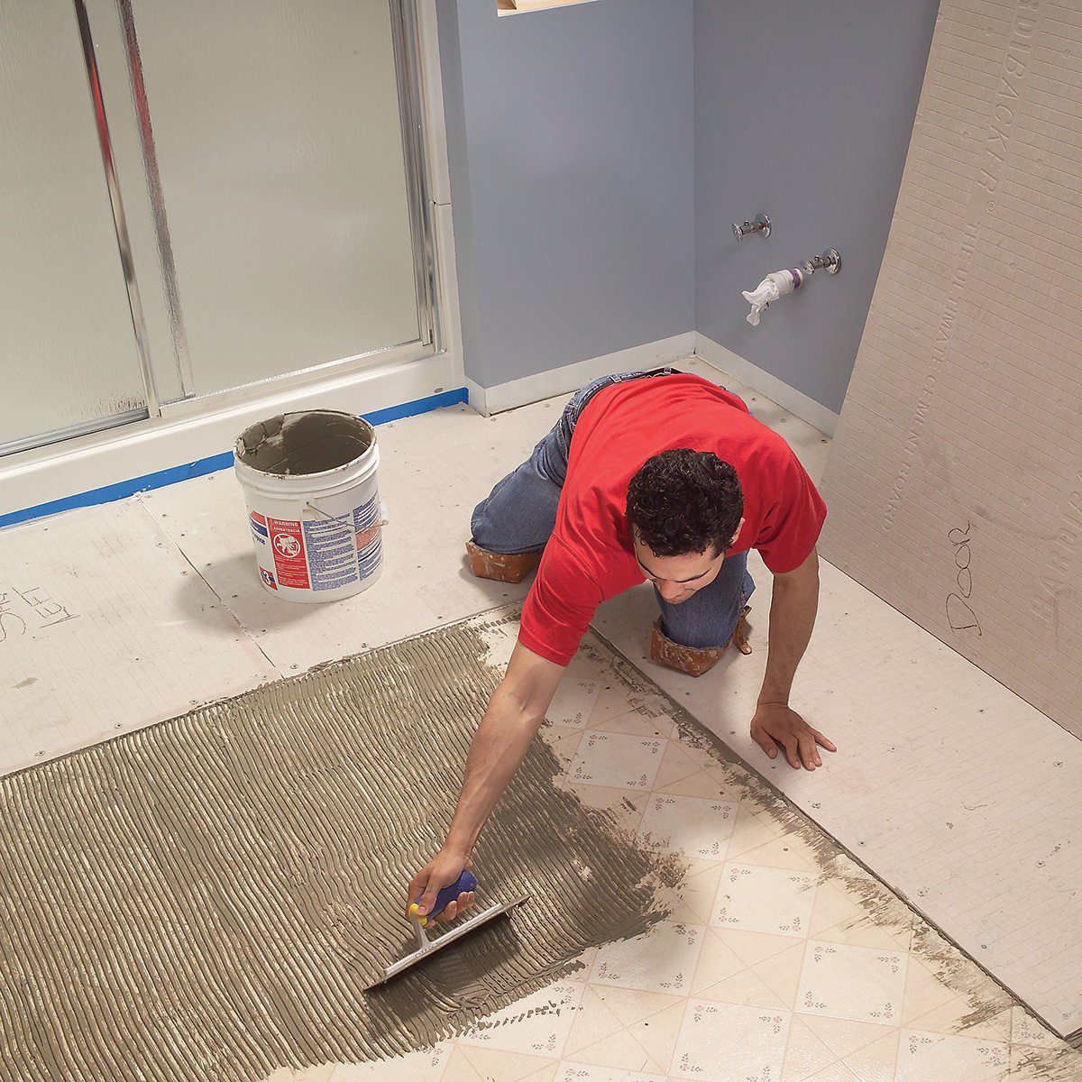 A person applies adhesive on a floor using a trowel, surrounded by a partially renovated bathroom featuring light blue walls and a bucket of adhesive.