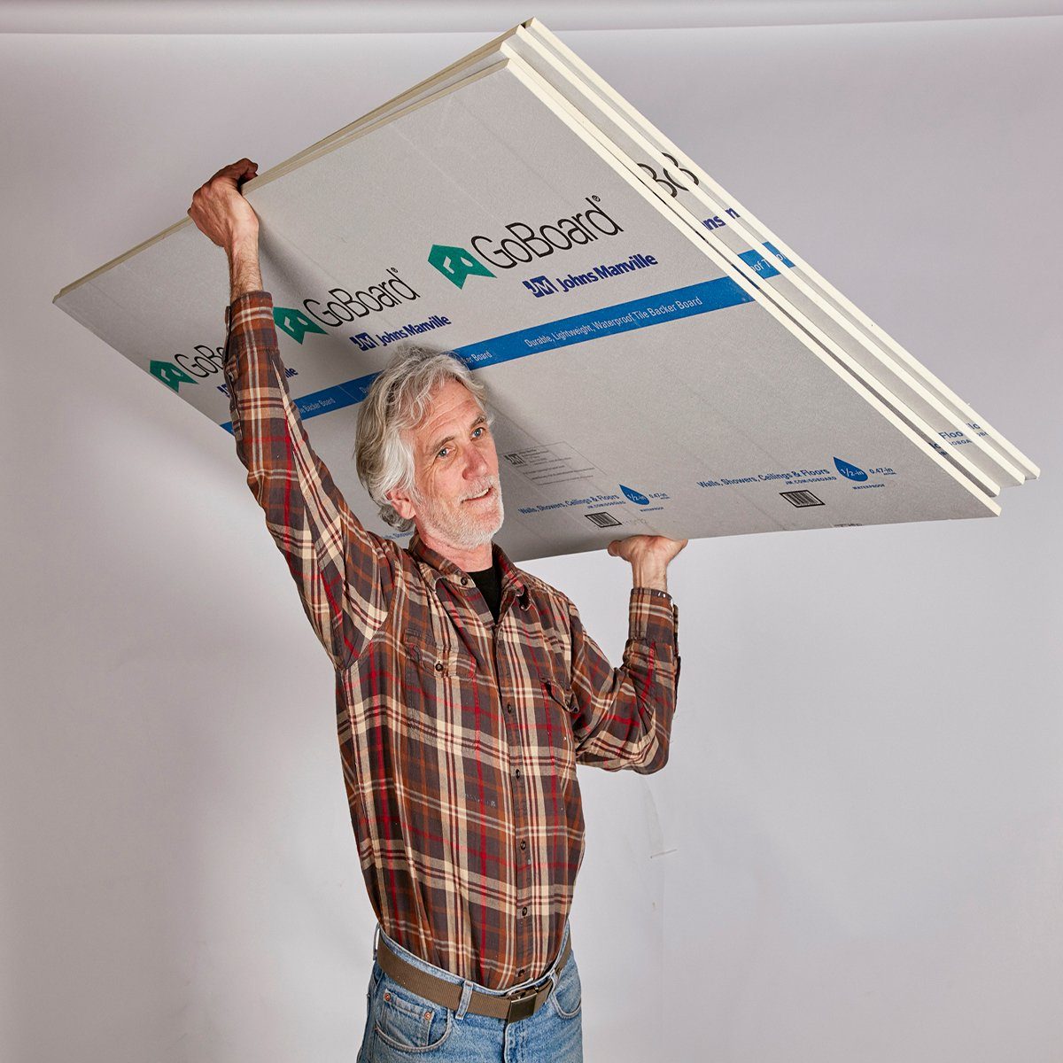 An elderly man is lifting large sheets of building material overhead, smiling. The backdrop is a simple, light-colored studio setting.