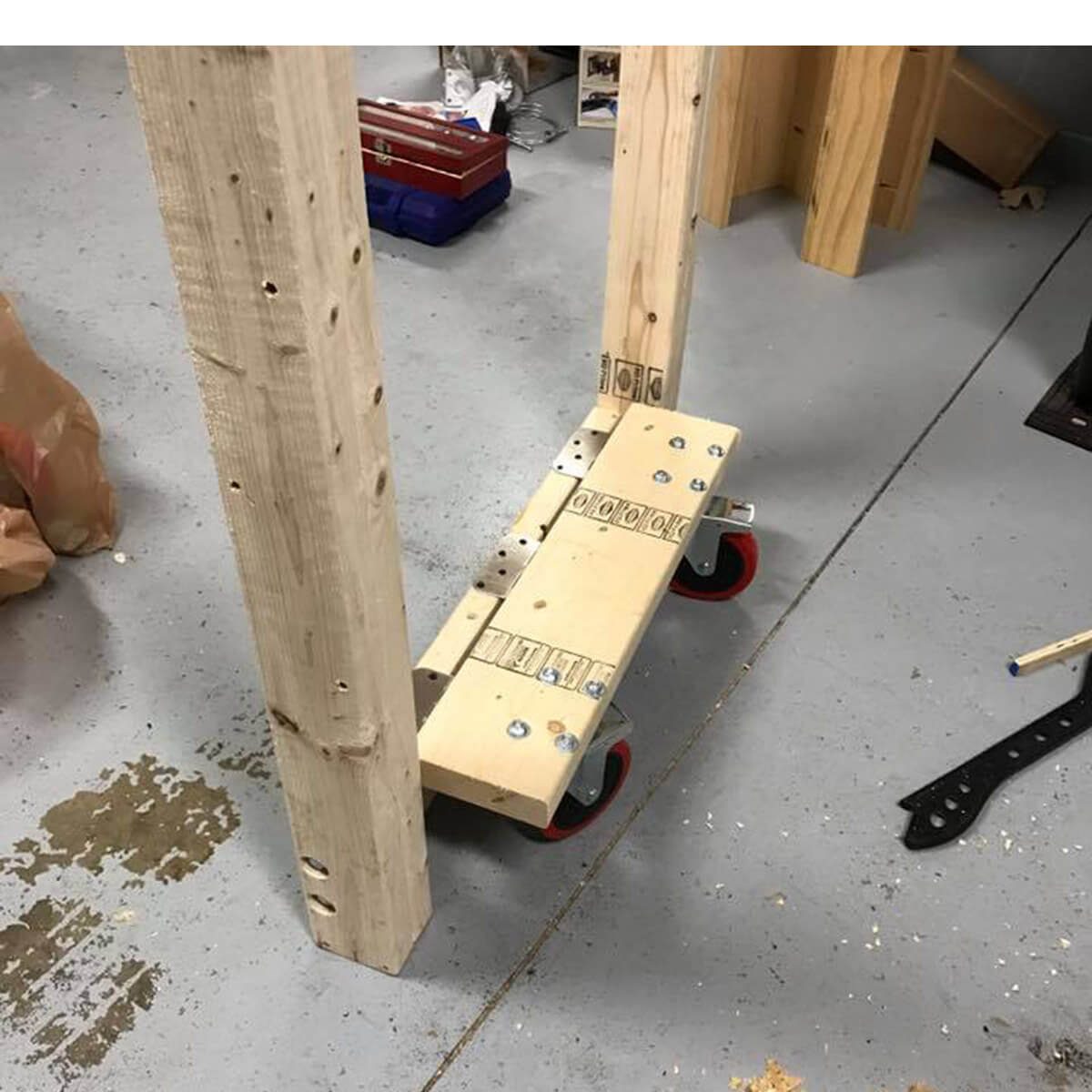 A wooden platform on wheels sits between two vertical wooden beams, resting on a concrete floor, surrounded by tools and scattered debris.