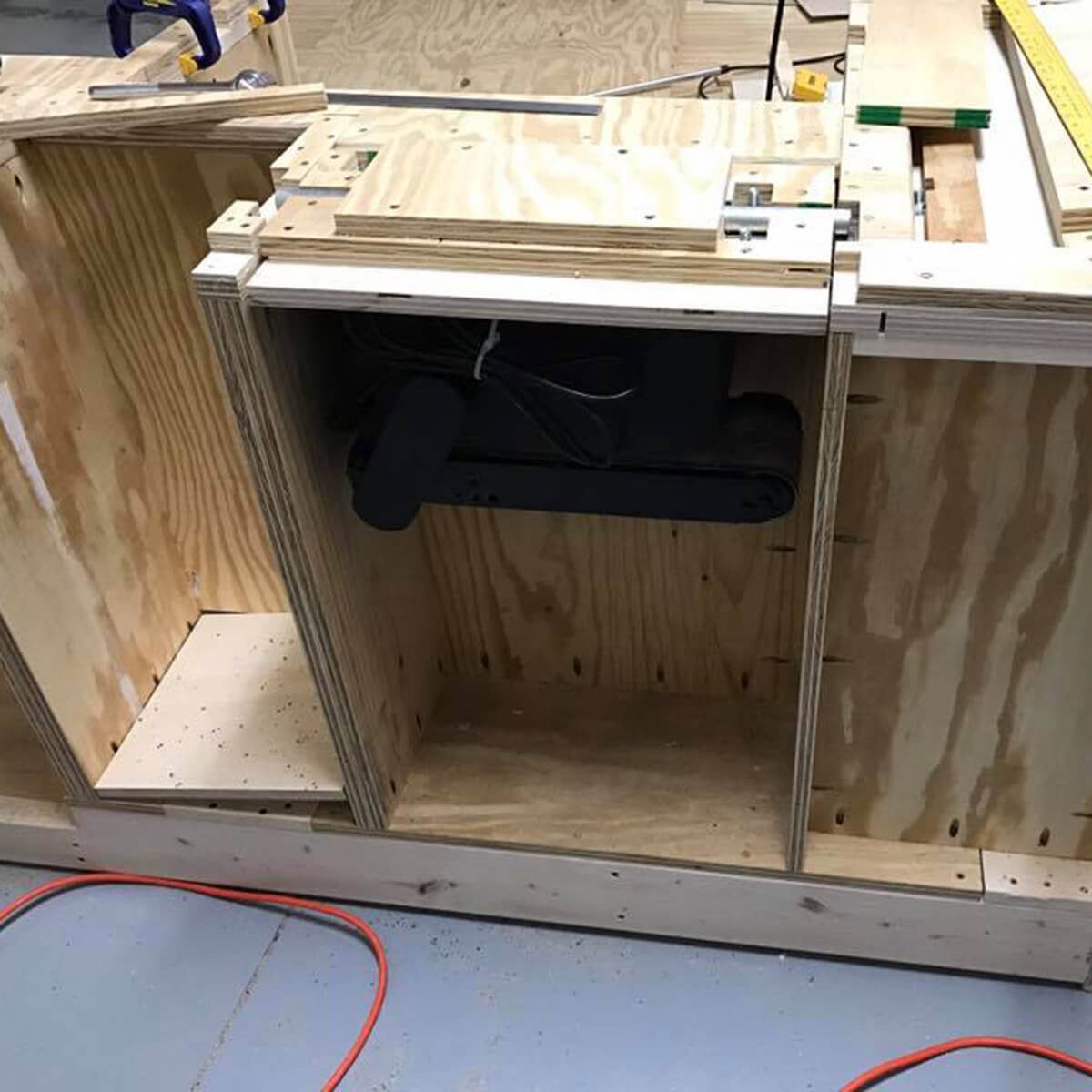 A wooden structure with compartments houses a motorized component inside. Power cords are visible on the floor in a workshop setting.