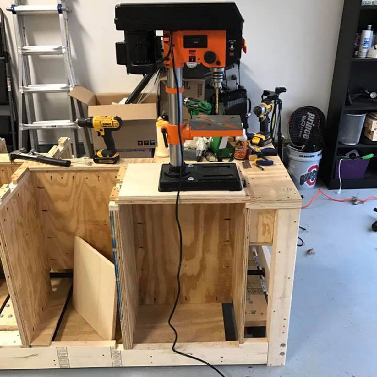 A drill press stands on a wooden workbench, surrounded by tools and partially assembled wooden boxes in a garage-like setting.