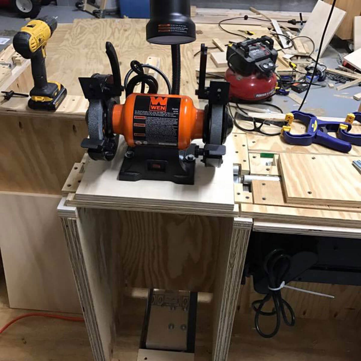 A bench grinder is set on a wooden worktable, illuminating the workspace with a lamp. Nearby, a cordless drill and various tools are scattered around.