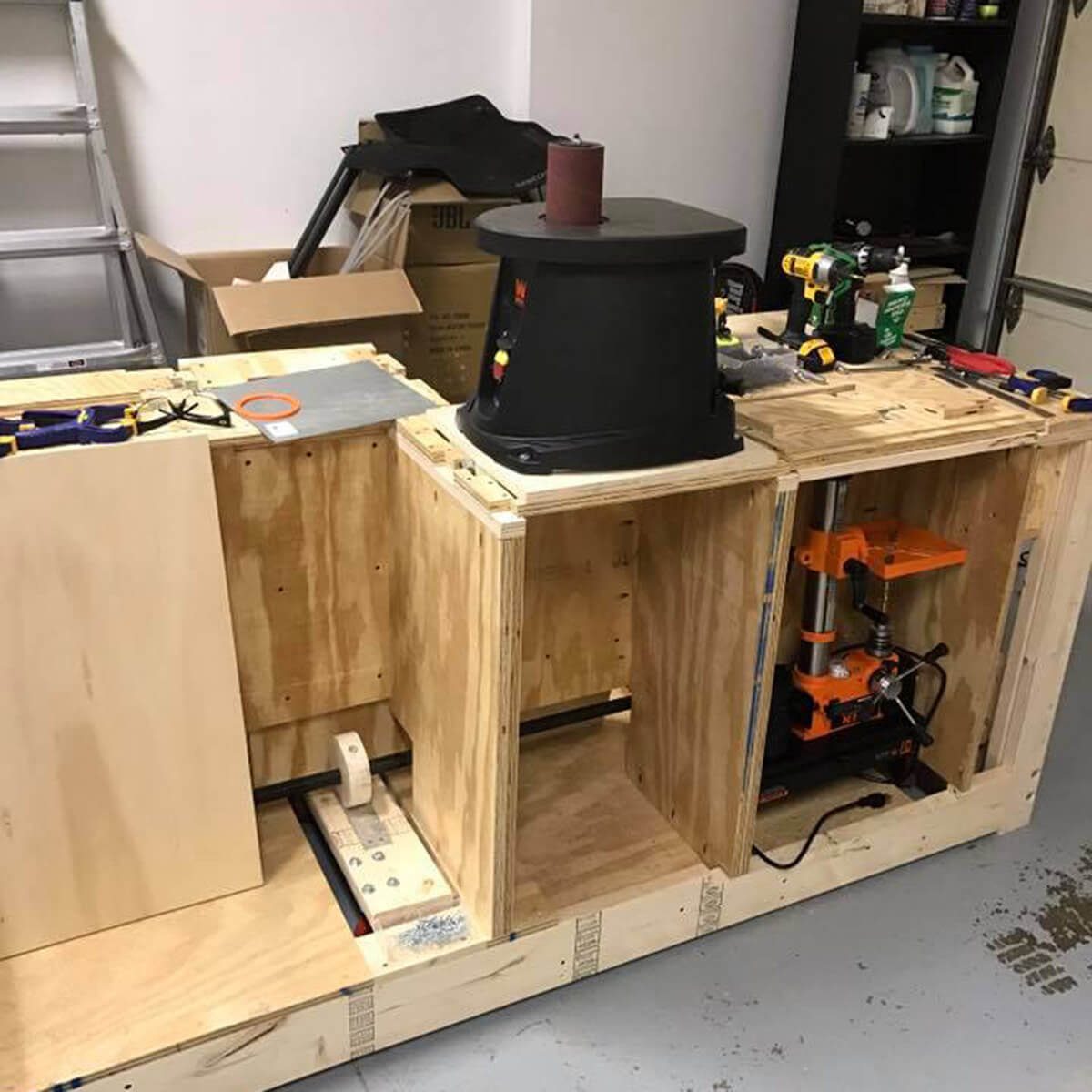A workbench features a black power tool atop wooden frames, surrounded by tools, boxes, and equipment, in a garage-like environment.