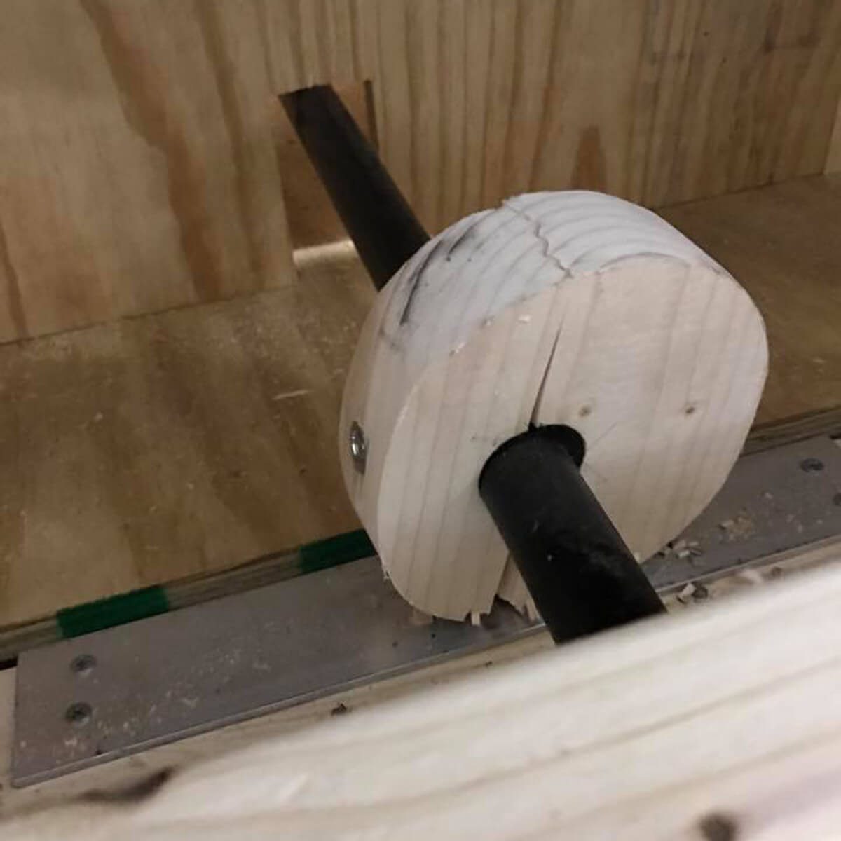 A wooden wheel attaches to a black rod, partially inside a wooden structure, with visible shavings on a metal track below.