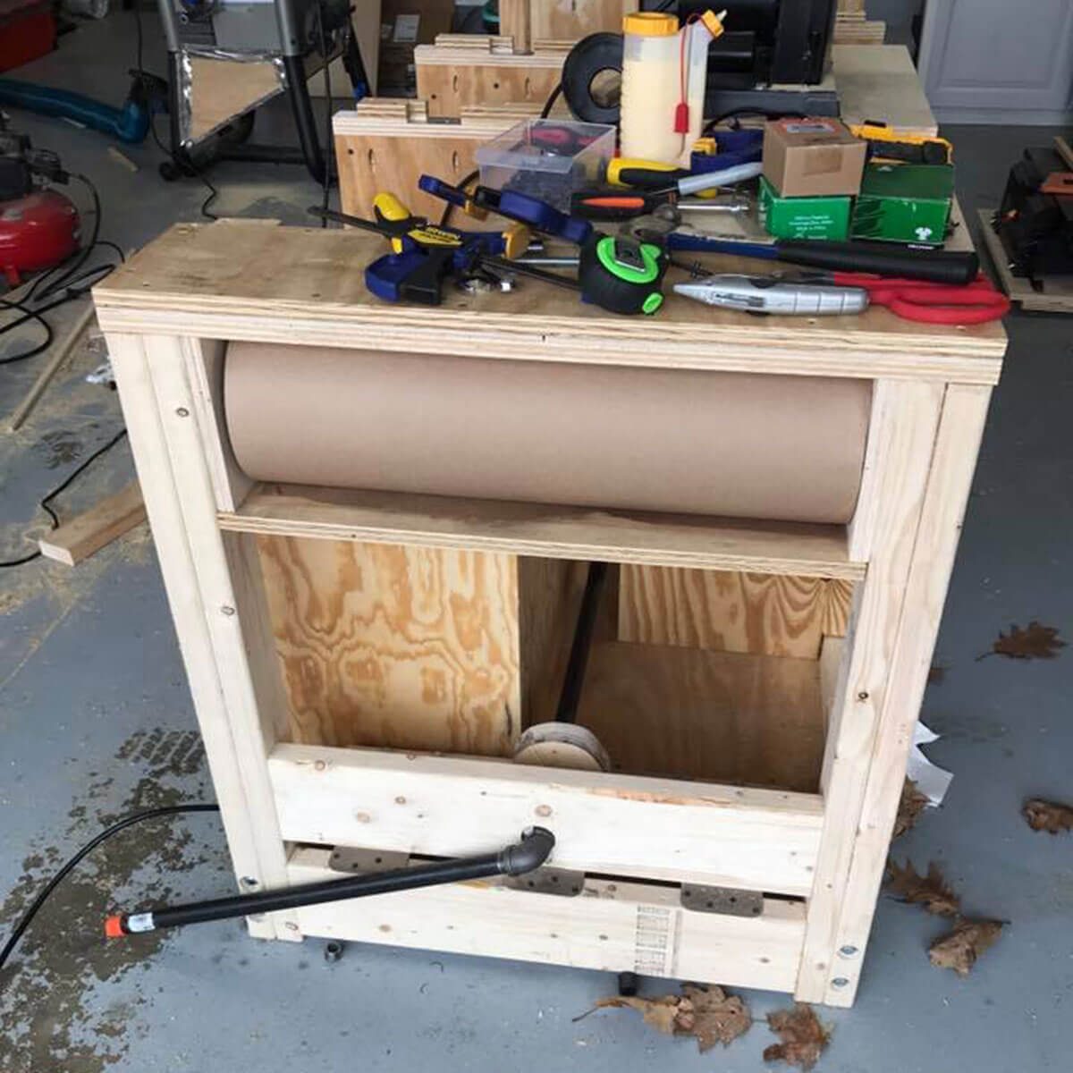 A wooden workbench displays various tools, with a rolled paper dispenser integrated beneath. The setting is a cluttered workshop with scattered materials.