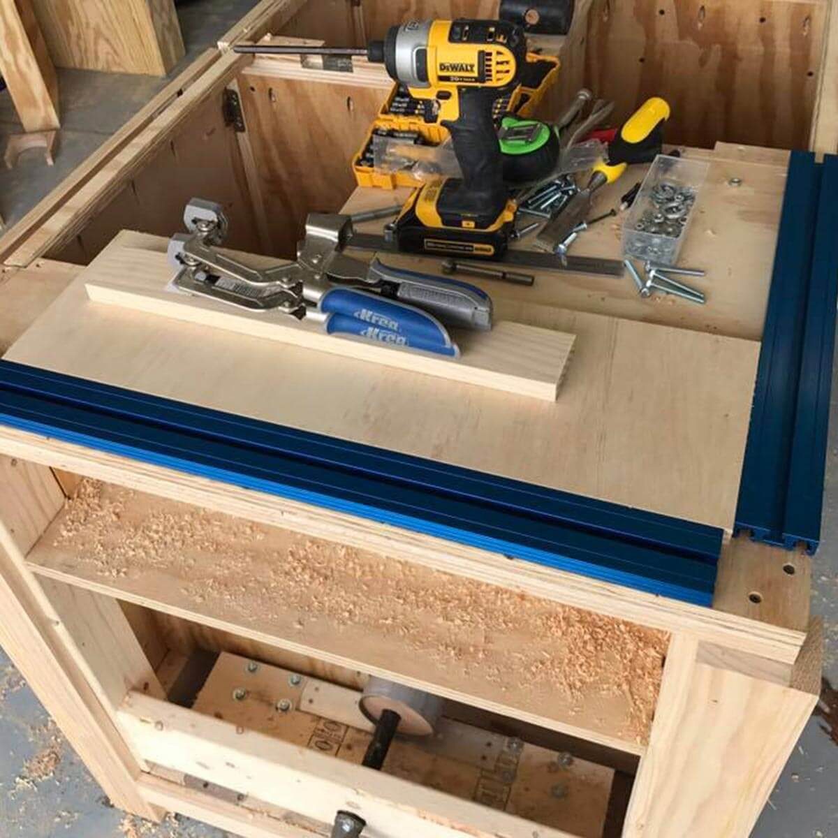 A workbench is filled with woodworking tools, including a drill and clamps, on a plywood surface, surrounded by wood shavings and unfinished wood.