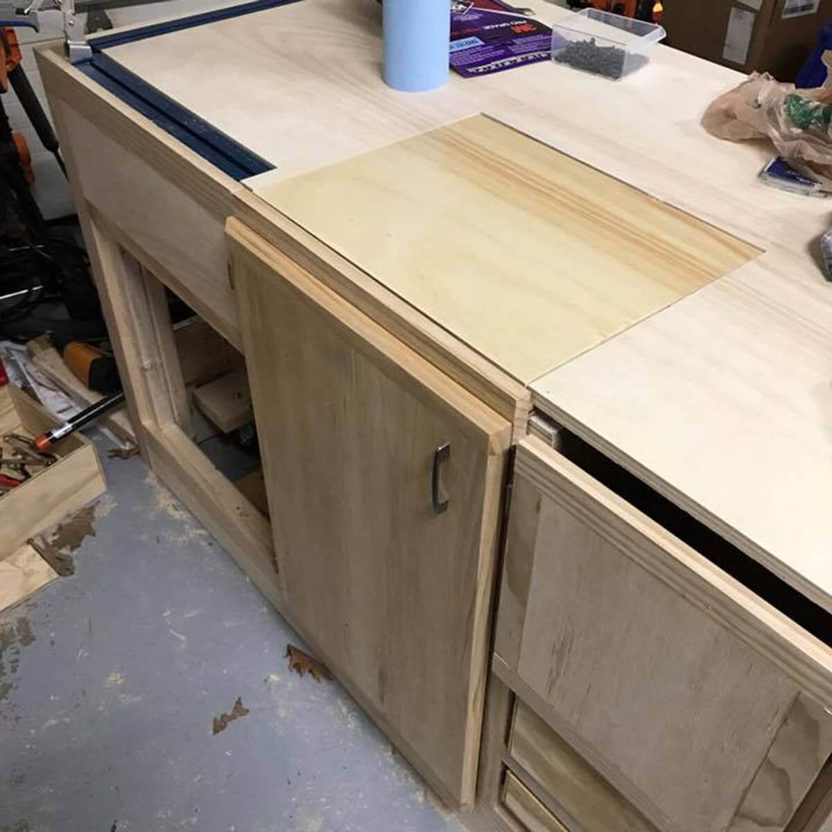 A wooden workbench with a cabinet door partially open; tools and materials are scattered around in a workshop setting.