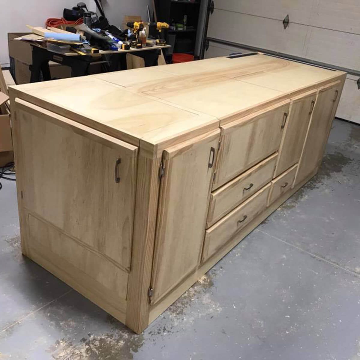 A large, unfinished wooden cabinet with drawers and doors is positioned in a garage, surrounded by tools and storage boxes.