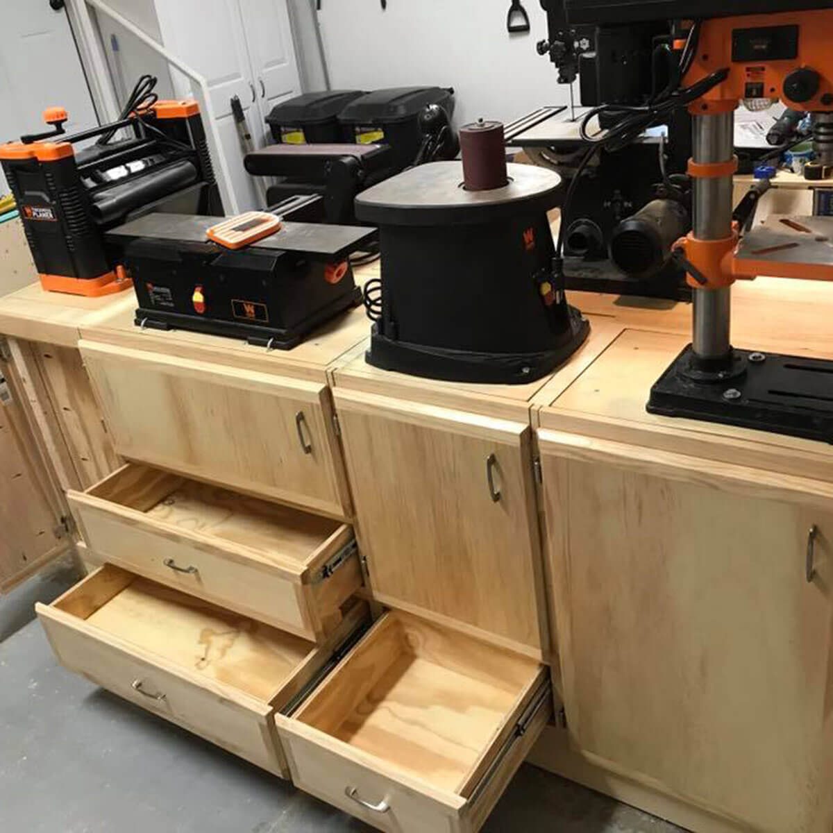 A wooden workbench displays various tools and machines on its surface, with multiple drawers partially opened, in a well-lit workshop environment.