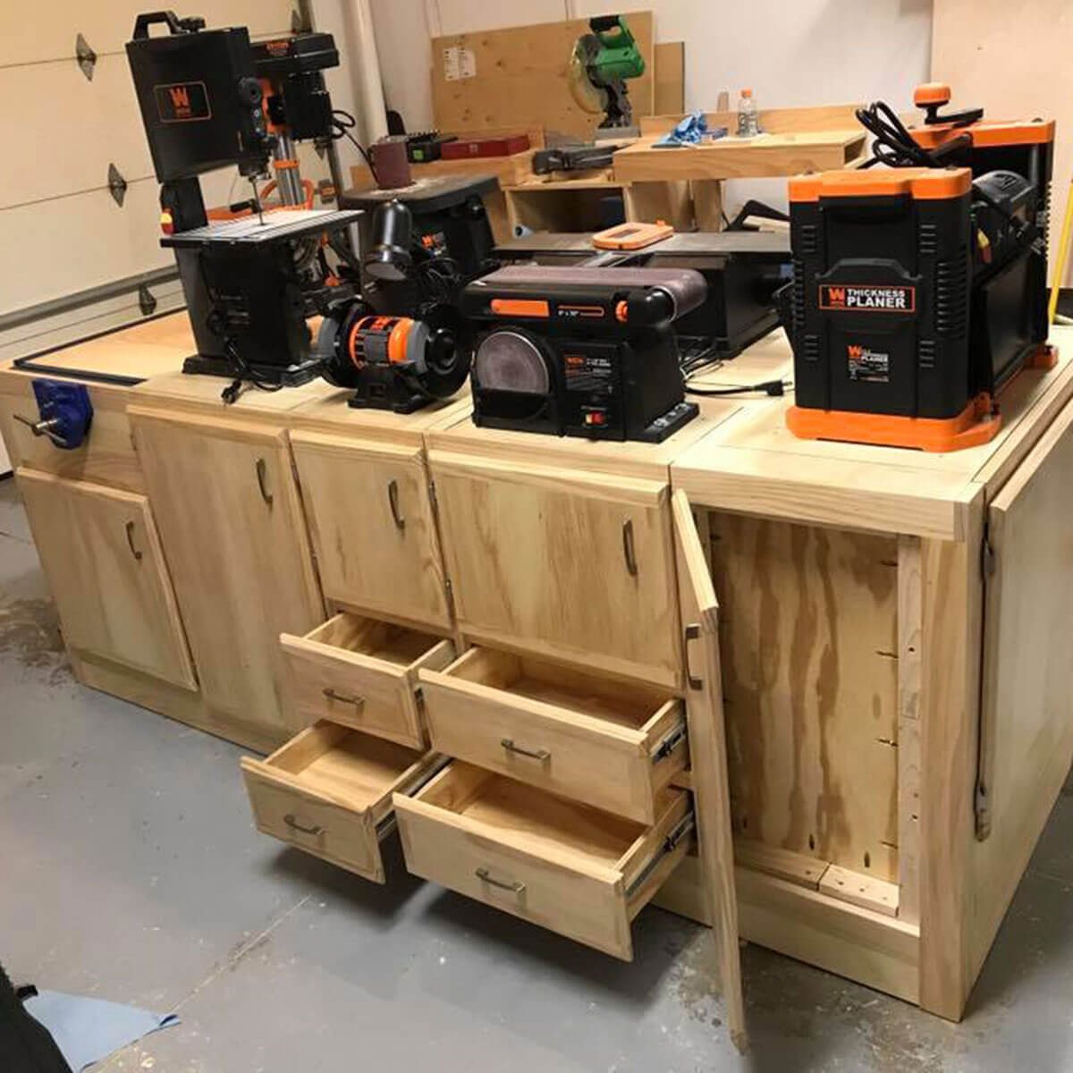 A wooden workbench holds various tools, including a planer and sander, with several open drawers revealing storage space, all set in a workshop environment.