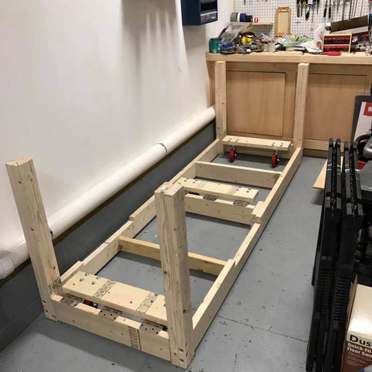 A wooden frame with wheels is being constructed in a workshop, surrounded by tools and workbenches against a clean, light-colored wall.