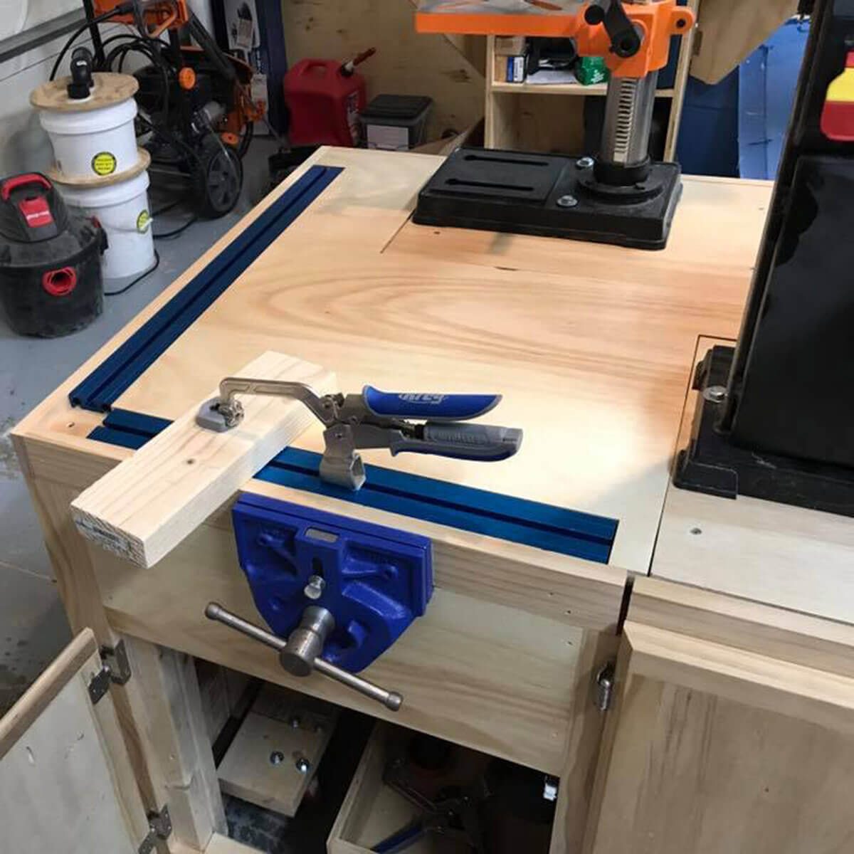 A wooden workbench holds a clamped piece of wood, with tools visible nearby in a garage workshop environment, including a vise and a drill press.