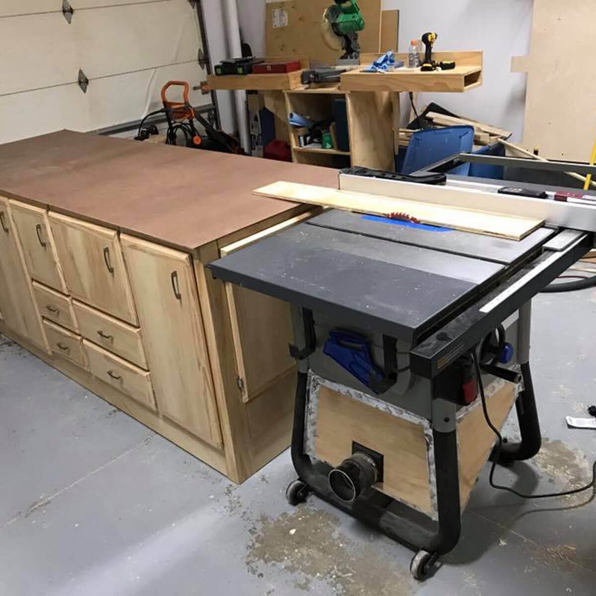 A wooden workbench with drawers stands beside a table saw, which is positioned to cut a piece of wood, in a garage workshop.