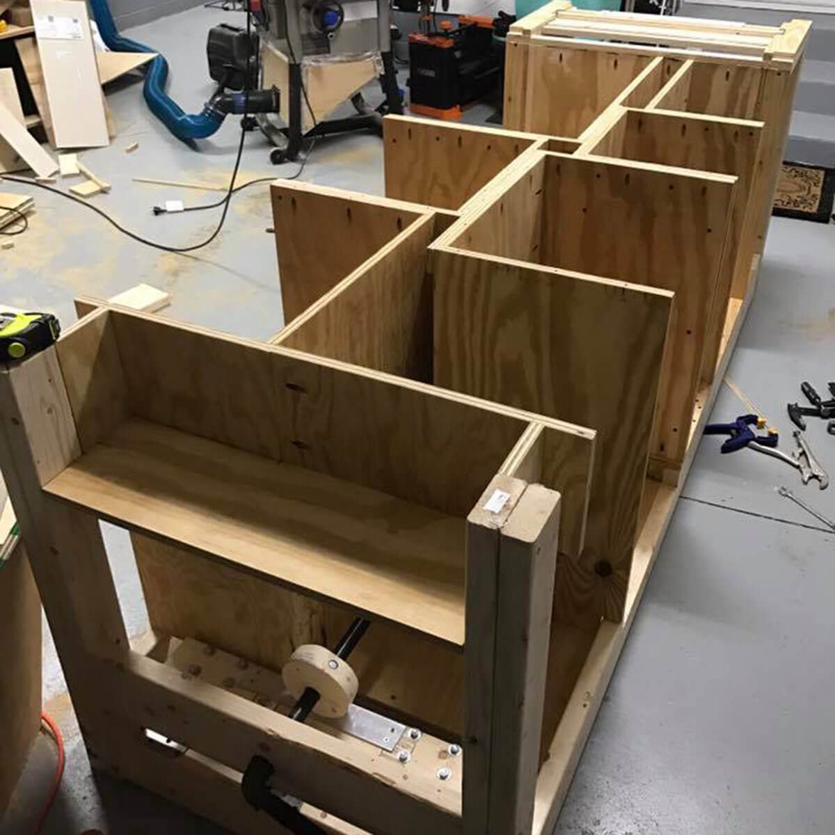A wooden structure with multiple compartments is being assembled in a workshop, surrounded by tools and materials, indicating an ongoing construction project.