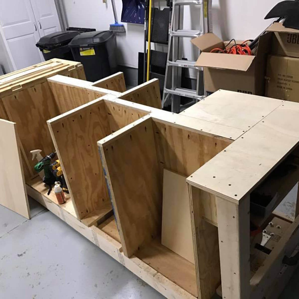 A wooden structure with compartments is partially assembled on a garage floor, surrounded by tools, boxes, and waste bins in a cluttered workspace.