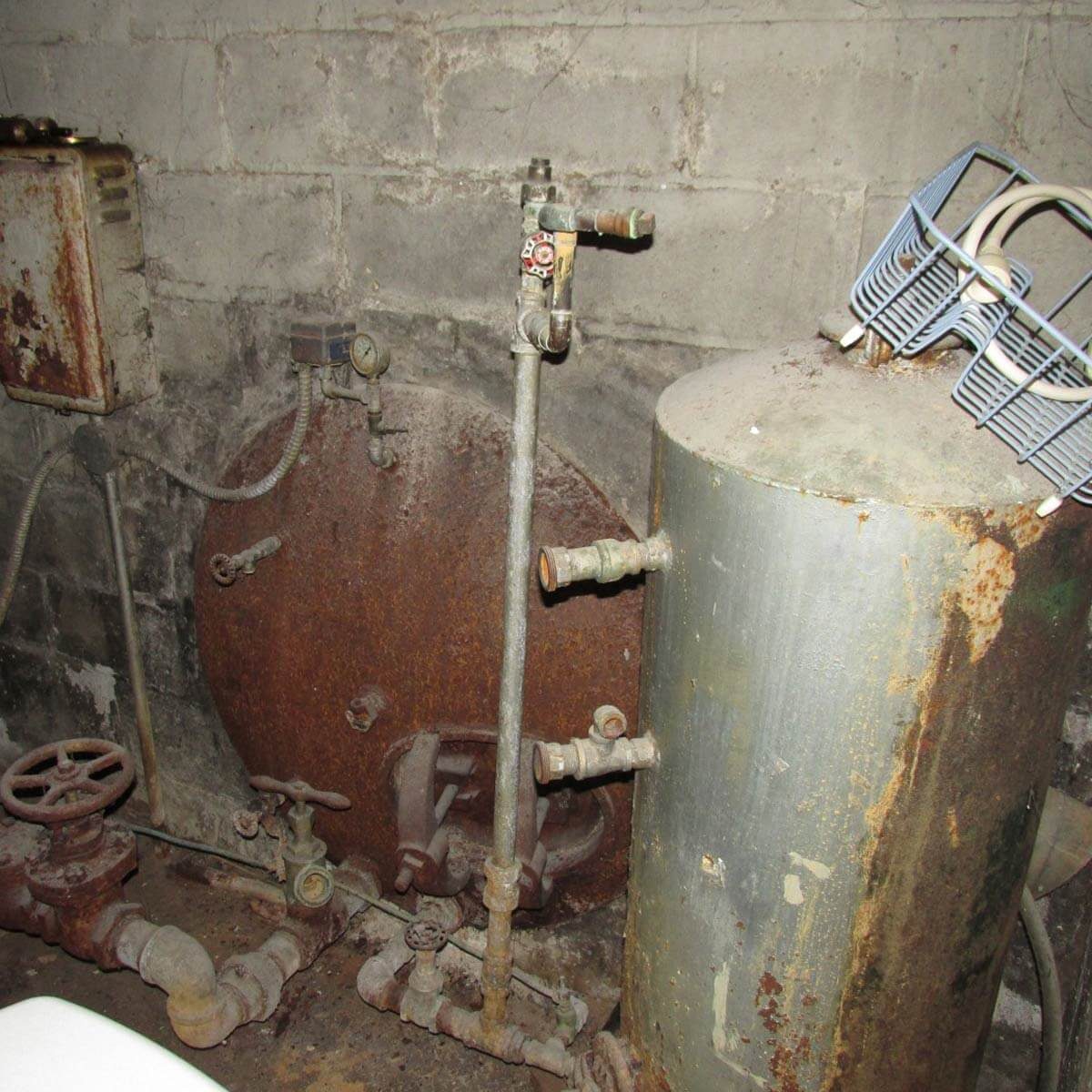 A rusty metal tank stands alongside aged pipes and valves in a dimly lit basement environment, with peeling walls and visible dust accumulation.