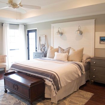 A neatly made bed with decorative pillows rests against a light-colored wall; two nightstands and a vintage trunk are positioned nearby in a cozy bedroom.