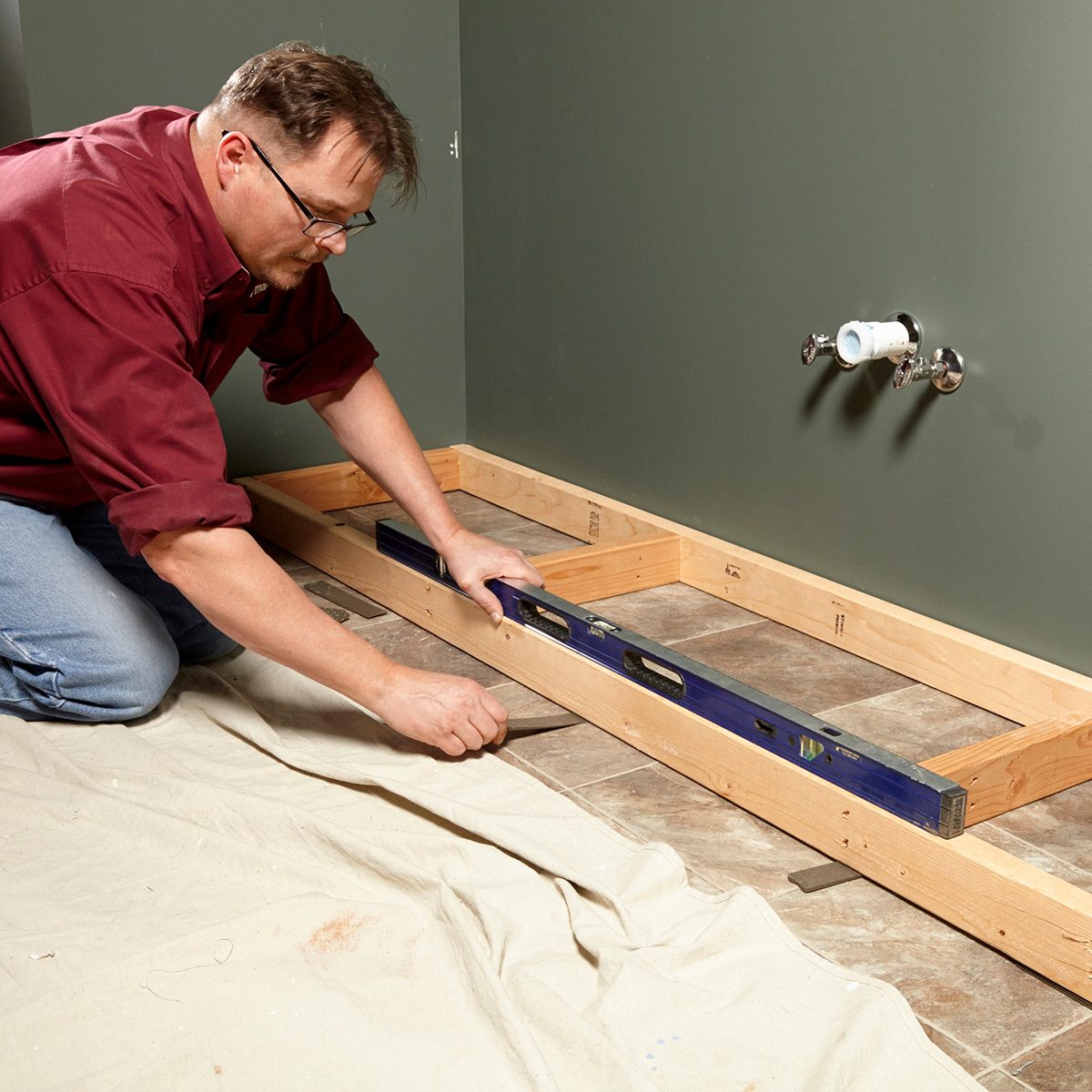 A man adjusts a level on wooden framing, ensuring it’s straight, while working on a tiled floor beside a green wall with plumbing fixtures.