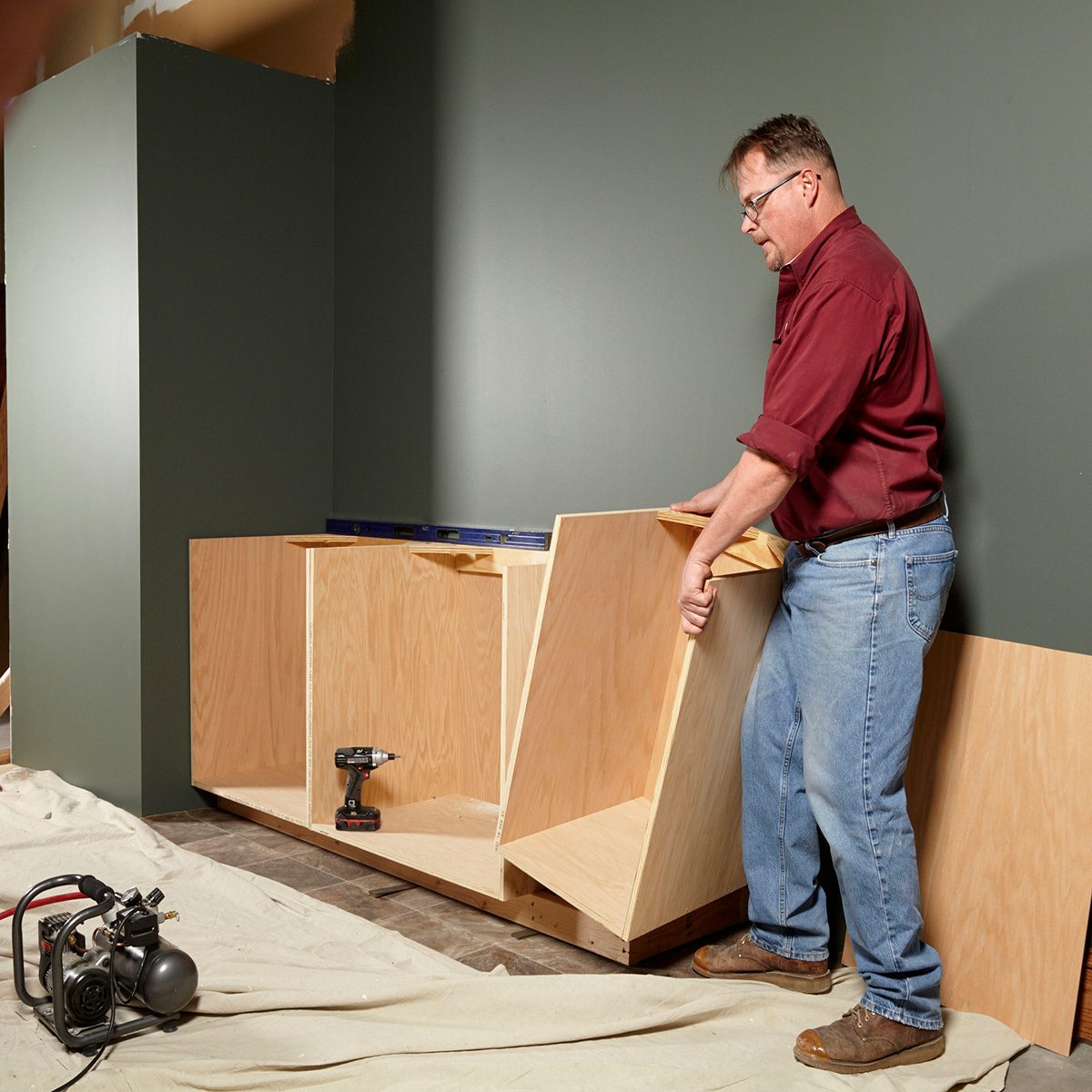 A man assembles wooden cabinetry in a room with dark green walls, surrounded by tools and a protective tarp on the floor.