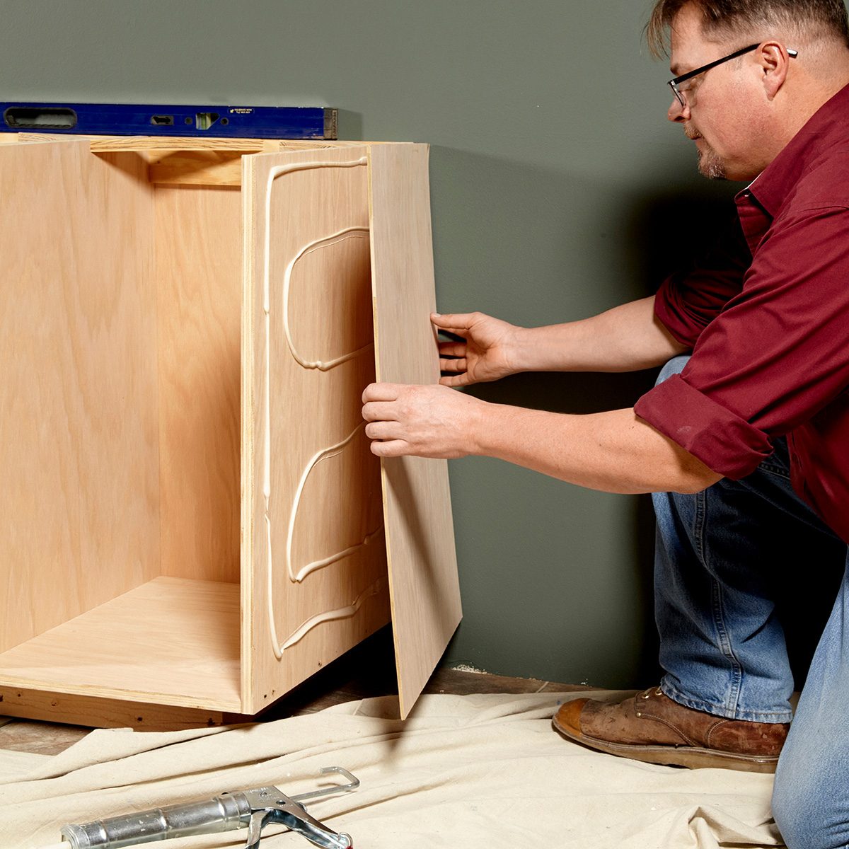A person is attaching a wooden panel to a cabinet while applying adhesive, with a level positioned above on a neutral-colored wall.
