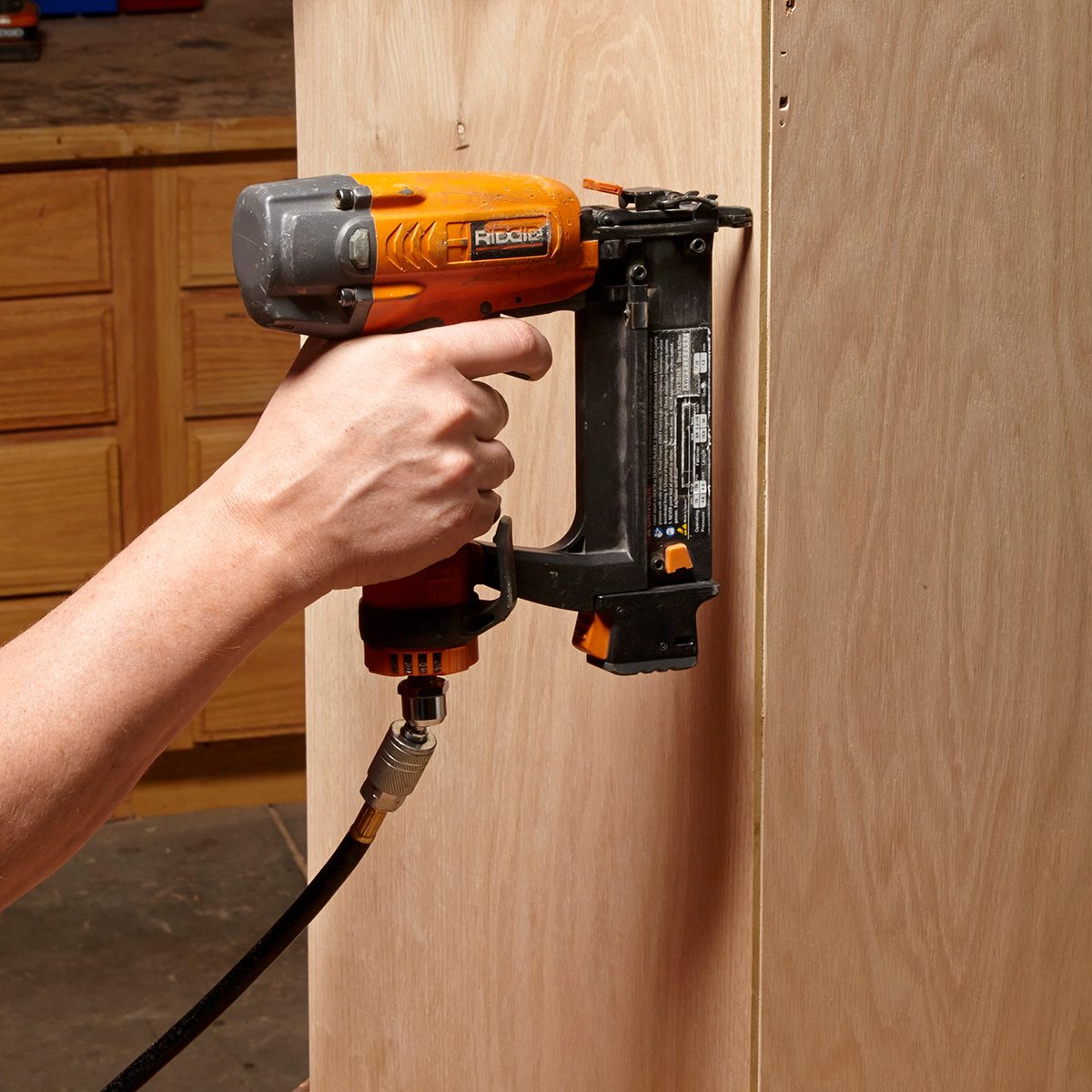 A hand holds a nail gun, driving nails into a wooden surface while surrounded by blurred cabinetry and tools in a workshop setting.