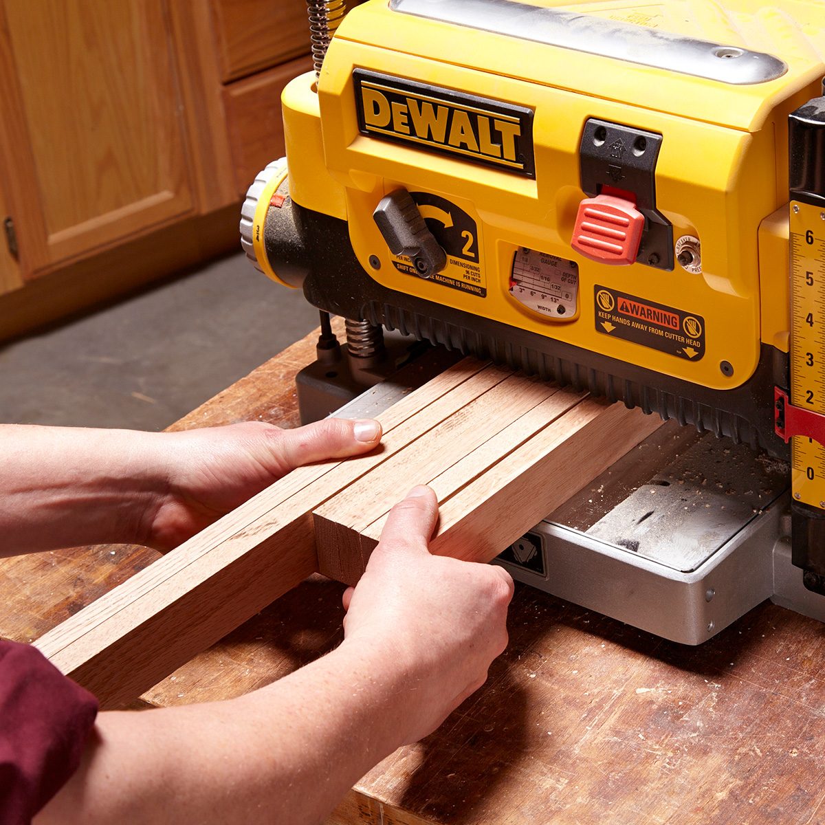 A person operates a yellow DEWALT planer, feeding wooden boards through it on a wooden workbench in a workshop setting.