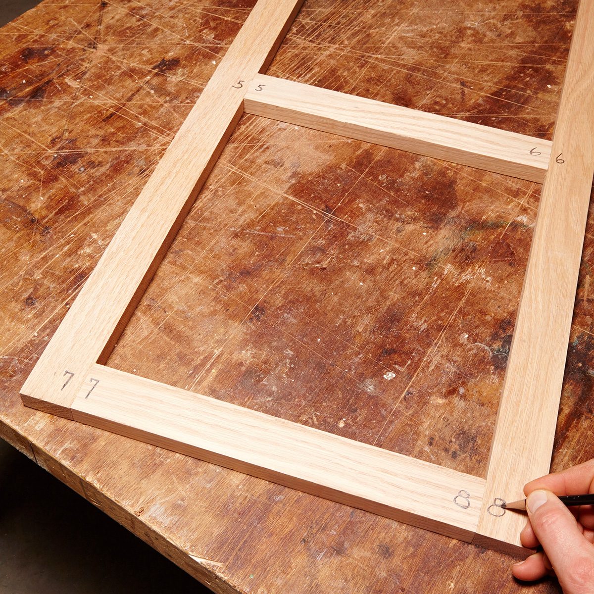 A wooden frame with marked dimensions is on a workbench. A hand is using a pencil to note measurements on the frame. The surface is stained.