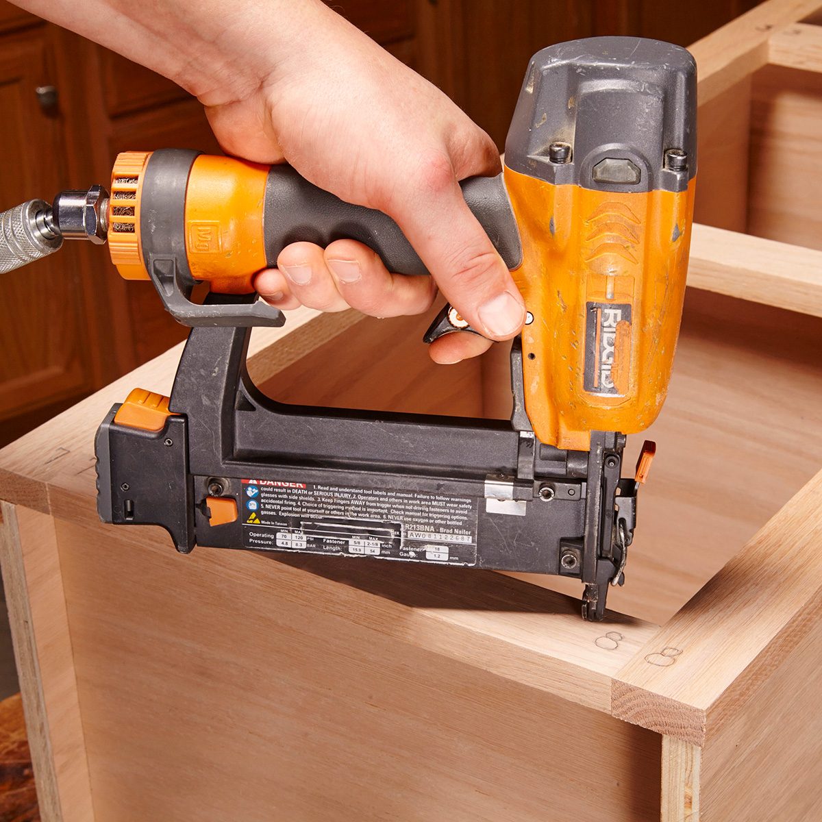 A person holds a nail gun, fastening wooden pieces at a corner in a workshop setting with wooden cabinets in the background.