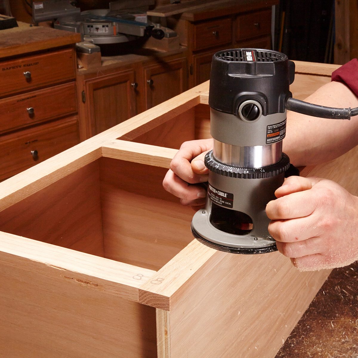 A person operates a router on a wooden box, shaping its edges, surrounded by a workshop with various tools and cabinets in the background.