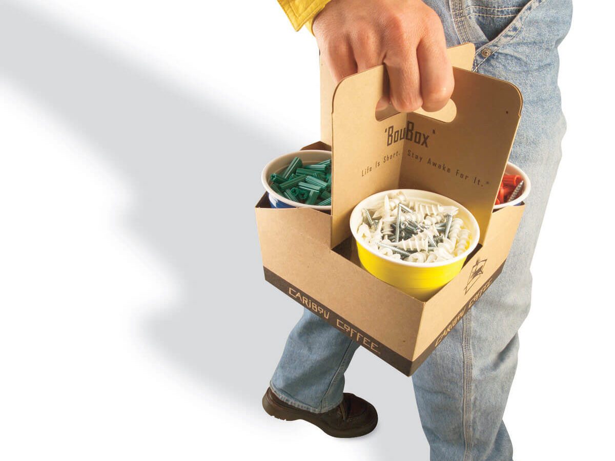 A person holds a cardboard caddy containing colored cups filled with various items while walking on a light background.