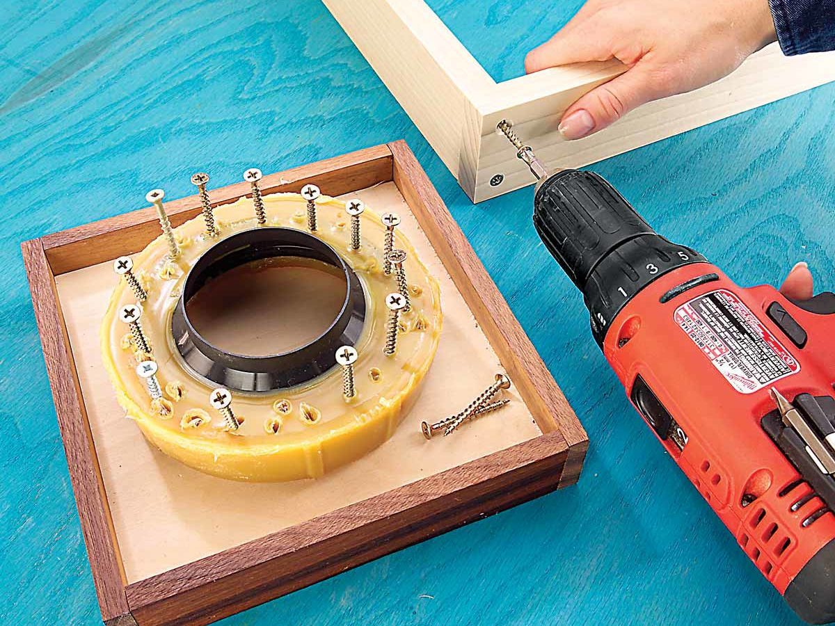 A person uses a power drill to insert screws into a wooden frame, with a circular jig containing screws resting on a blue surface.