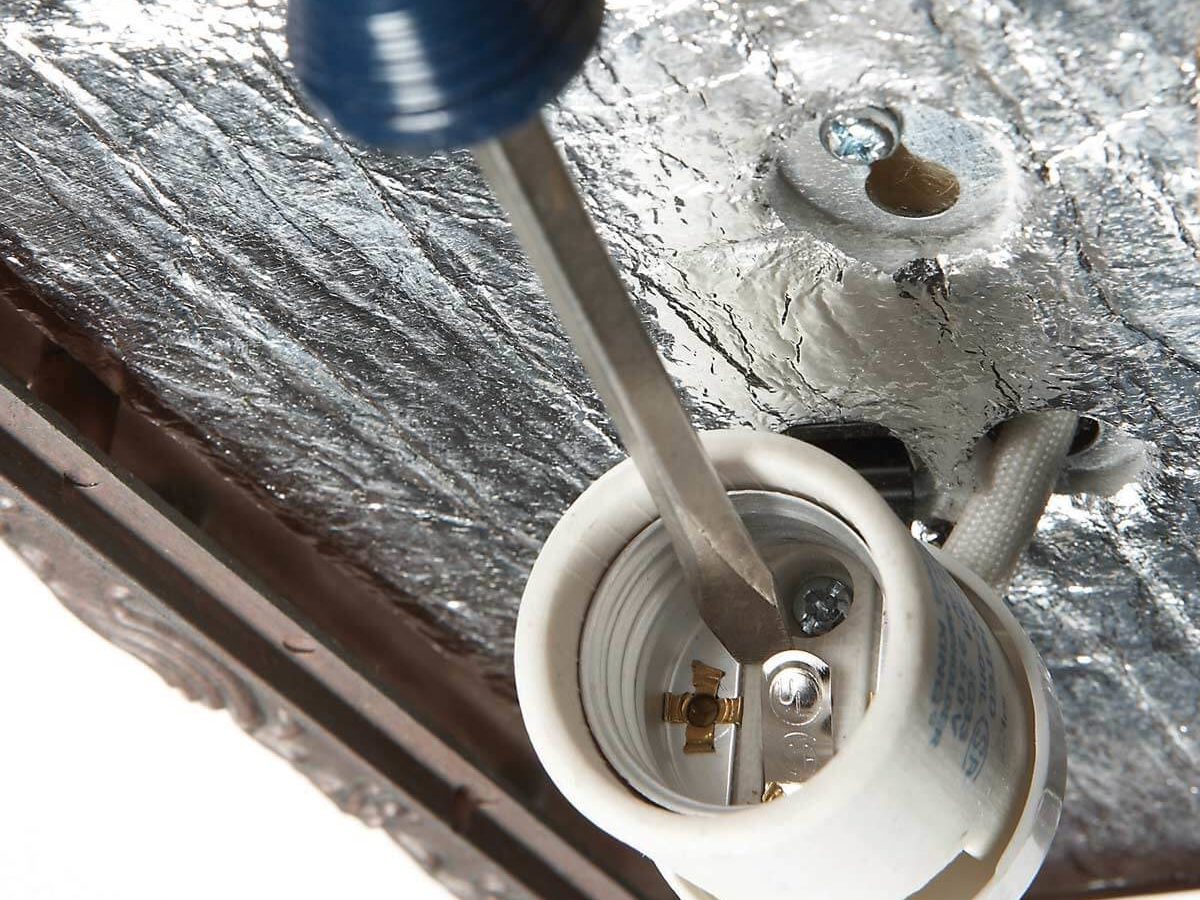 A screwdriver is inserted into a light socket to tighten connections, with a reflective surface above and structural framing visible around the scene.