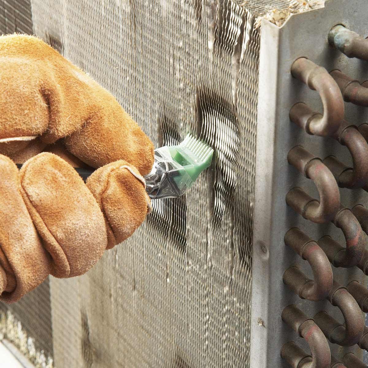 A gloved hand holds a cleaning tool, brushing the ribbed surface of a metal coil, with copper pipes visible on the side, indicating maintenance work.