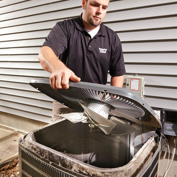 A technician lifts the cover of an air conditioning unit, inspecting its internal components, with a house's exterior siding visible in the background.