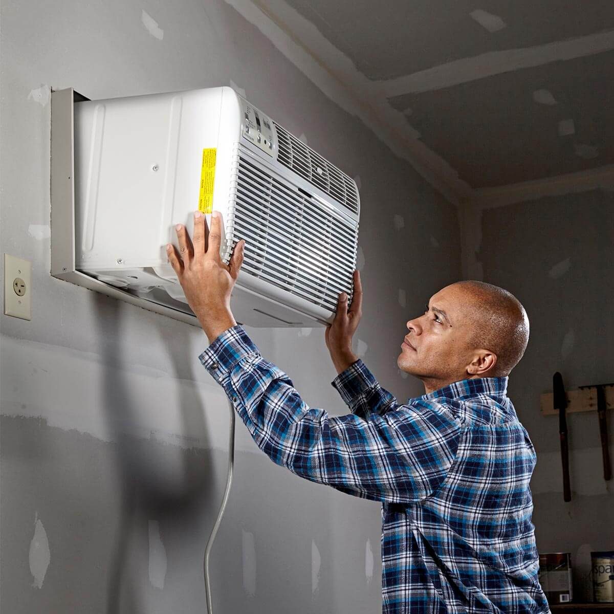 A man installs a window air conditioning unit on a wall in a room with unfinished drywall, tools, and equipment nearby.