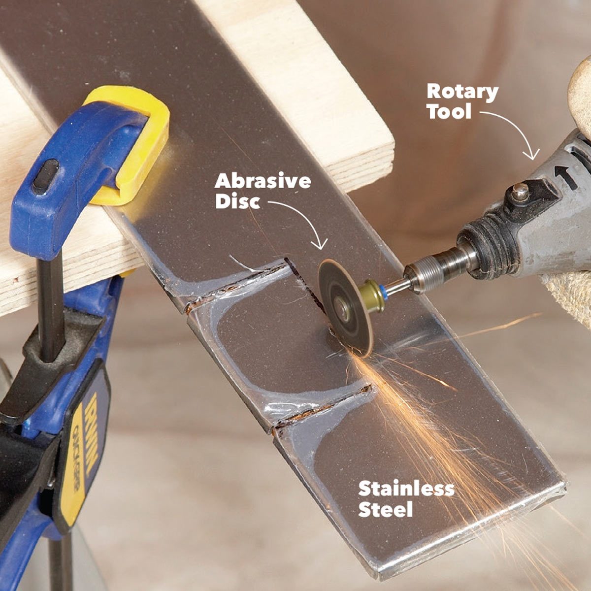 A rotary tool cuts stainless steel with an abrasive disc, producing sparks, while the metal is secured by a blue and yellow clamp on a wooden surface.