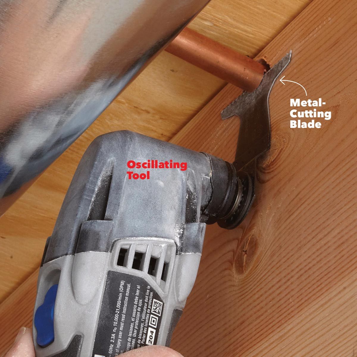 An oscillating tool equipped with a metal-cutting blade is cutting through wood near a copper pipe in a workshop environment.