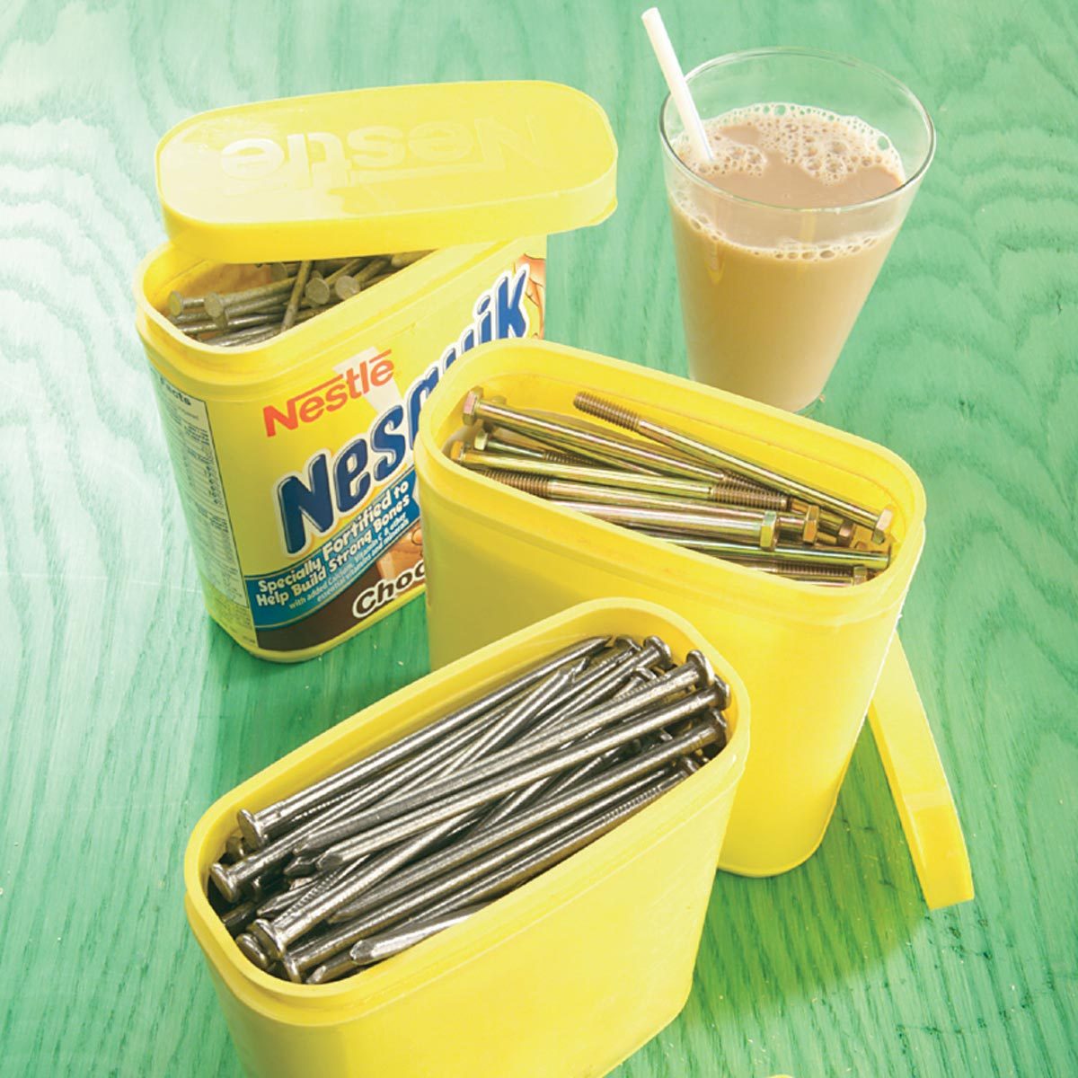 Two yellow containers filled with metal screws and bolts sit beside a glass of chocolate milk on a green wooden surface.